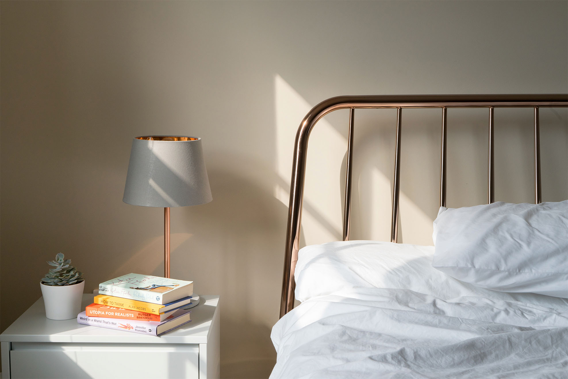 a bed mate up with white sheets with books and a fake plant on the bedside table